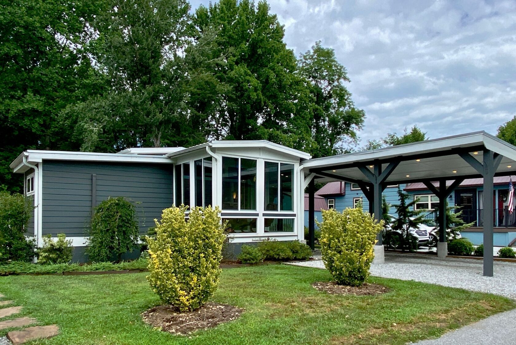 Modern gray house on Just Wandering Lane with large windows, carport, green lawn, shrubs, and blue sky in the background.