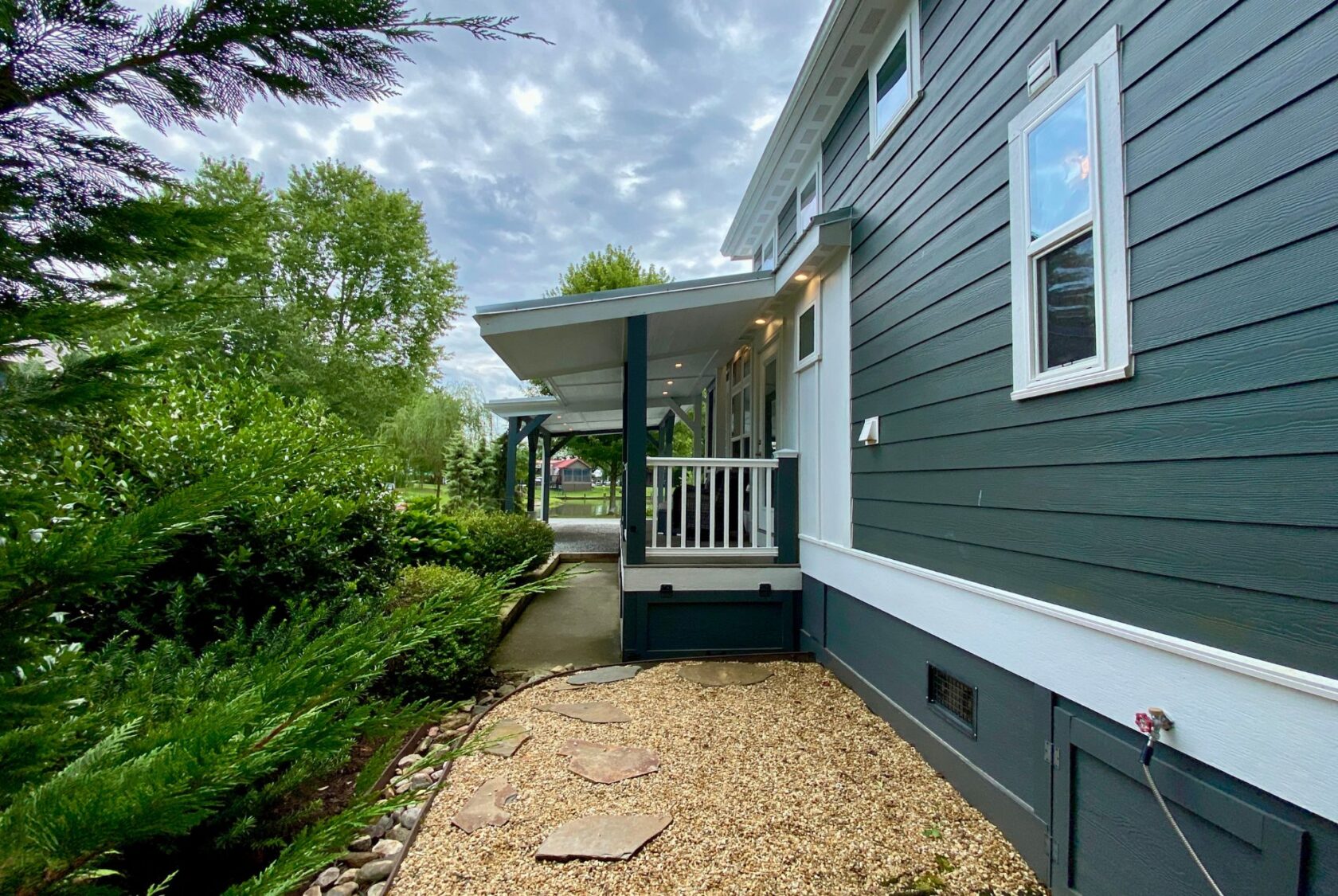 Side view of a dark green house on Just Wandering Lane, featuring a porch, gravel path, and lush plants under cloudy skies.