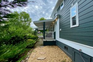 Side view of a dark green house on Just Wandering Lane, featuring a porch, gravel path, and lush plants under cloudy skies.