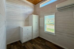 Small, bright room on Breezy Meadow Lane with white wood walls, dresser, wardrobe, window, and wall-mounted air conditioner.