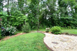 A backyard garden on Just Wandering Lane with green grass, blooming bushes, and a gravel path lined by trees.