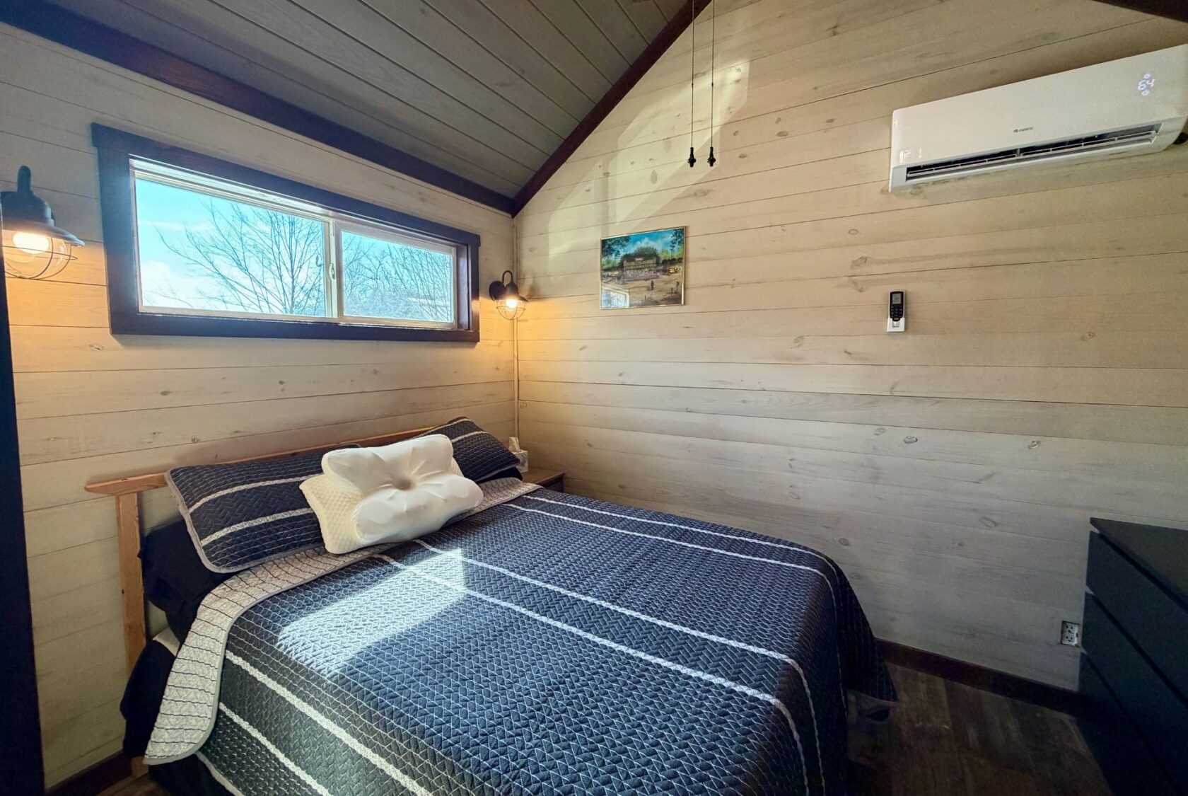 Cozy bedroom on Rest Awhile Lane with wood walls, double bed, two windows, wall art, and air conditioner above.
