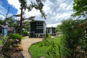 A tiny house with dark siding sits on Just Wandering Lane, surrounded by flowers and a lush green yard under a cloudy sky.