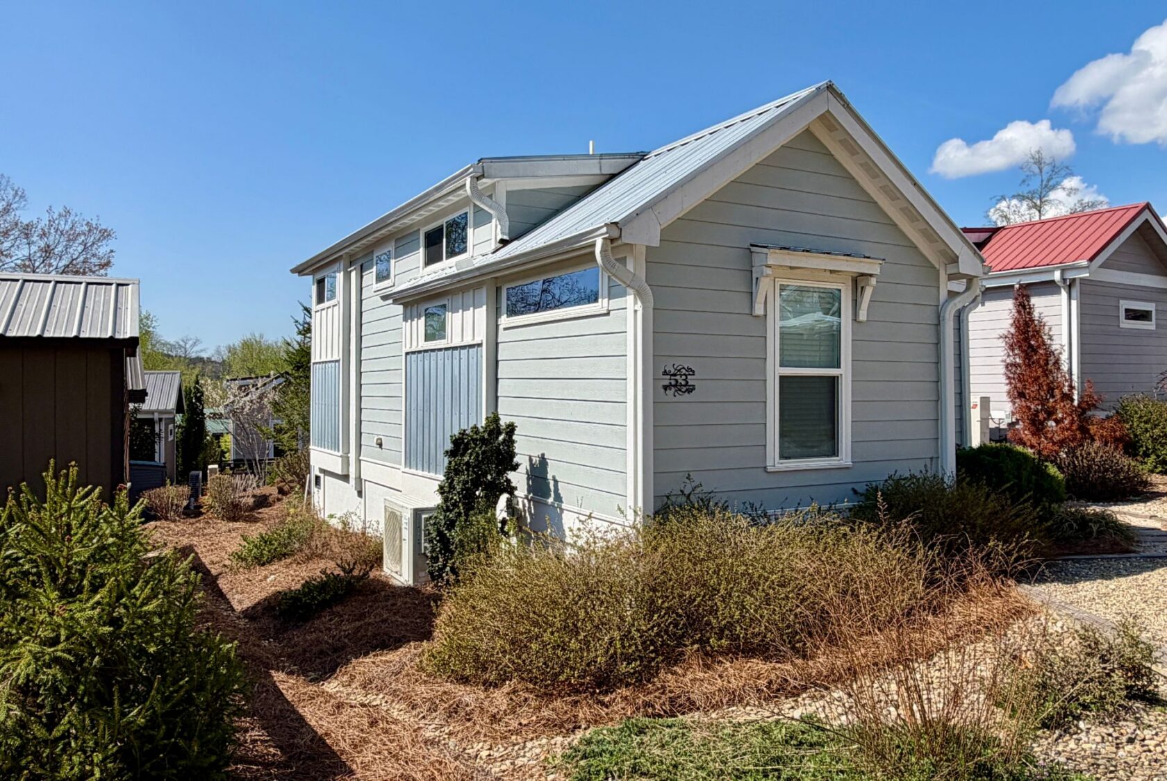 Light blue tiny house with white trim on Highland Hill Lane, surrounded by bushes and gravel pathways under a clear sky.