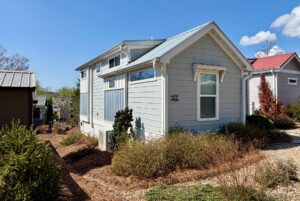 Light blue tiny house with white trim on Highland Hill Lane, surrounded by bushes and gravel pathways under a clear sky.