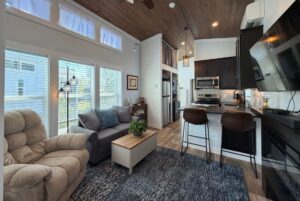 Cozy modern living room and kitchen on Meandering Lane with large windows, brown furniture, and wood ceiling accents.