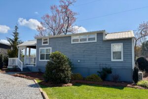 Charming light blue tiny house with white trim at 47 Drifting Brook, featuring a porch and garden under a blue sky.