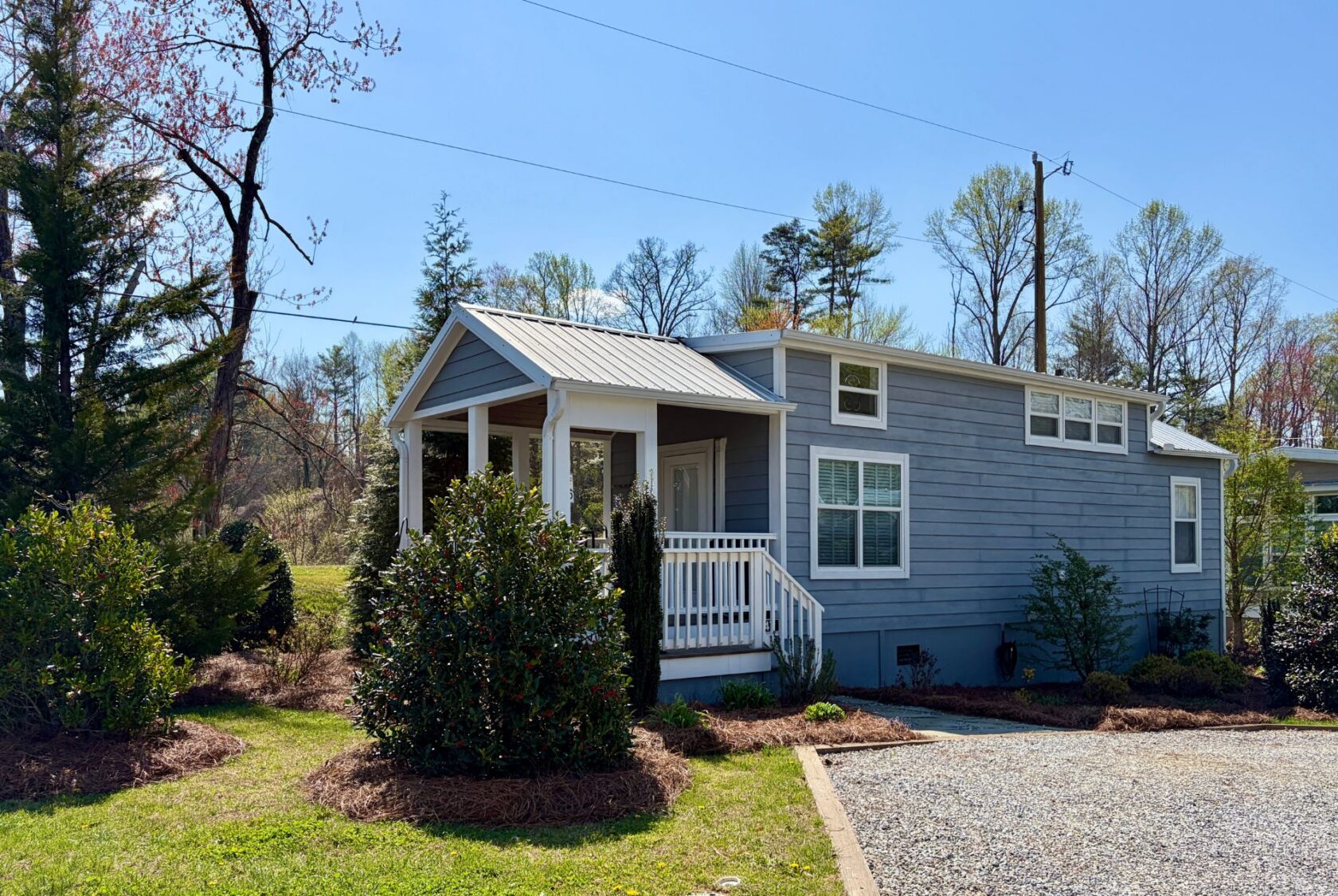 A small blue tiny house with a porch, white trim, and gravel drive at 47 Drifting Brook, surrounded by trees and bushes.