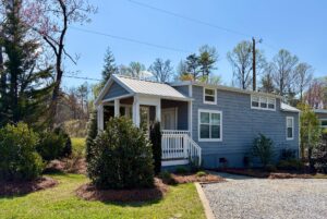 A small blue tiny house with a porch, white trim, and gravel drive at 47 Drifting Brook, surrounded by trees and bushes.