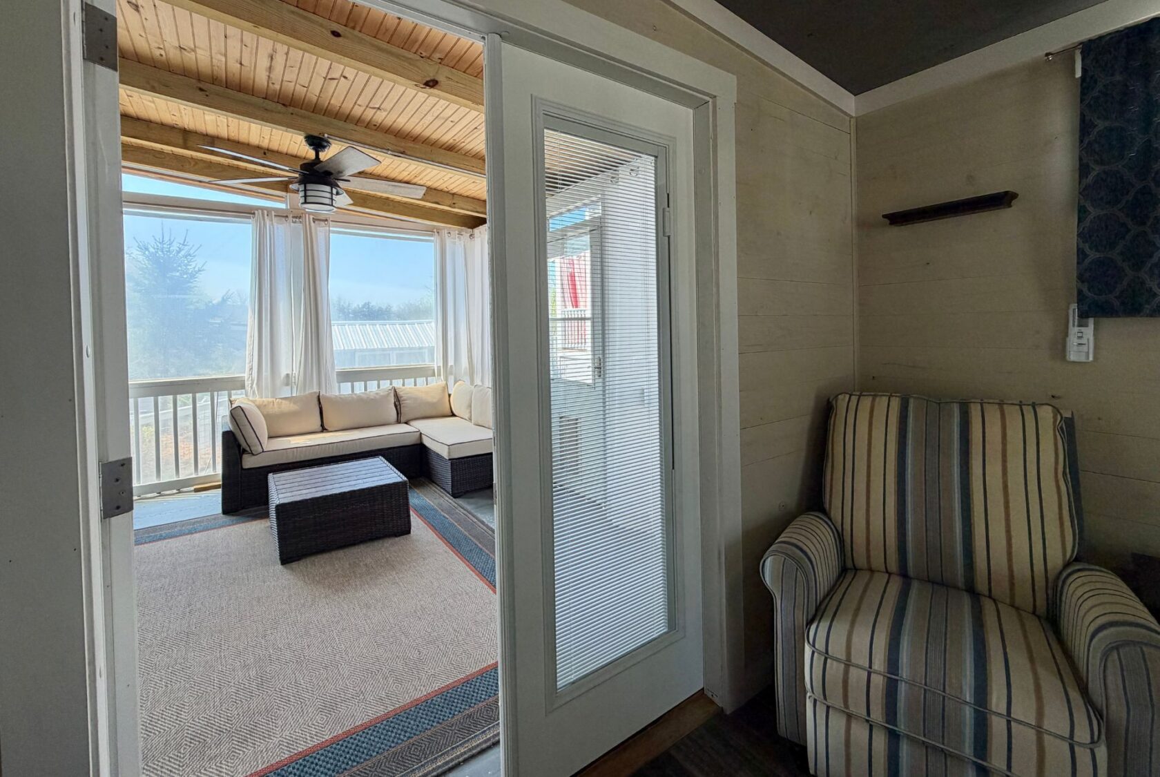 A cozy sunroom at 53 Highland Hill with a sectional sofa, wicker table, and striped armchair, viewed through glass doors.