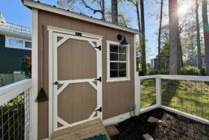 Charming small brown shed with white-trimmed door and window at 106 Meandering Lane, sunlight filtering through trees.