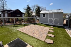Fenced backyard on Highland Hill Lane with artificial grass, stone path, paver patio, and gray shed under a clear sky.