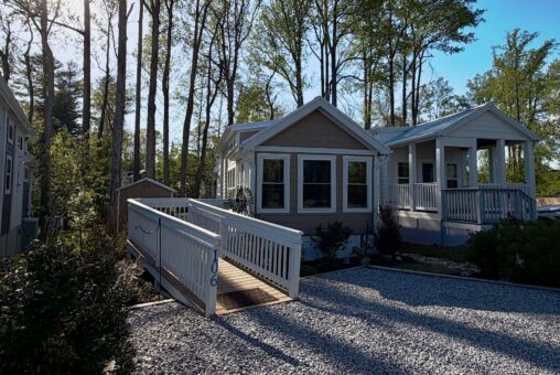 Charming small beige house at 106 Meandering Lane with a ramp, porch, and gravel yard nestled among trees in bright sunlight.