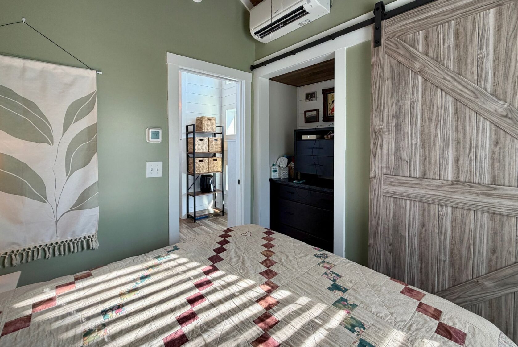 Sunlit bedroom at 106 Meandering Lane with a patterned quilt, green walls, barn door, and a view into shelves and baskets.
