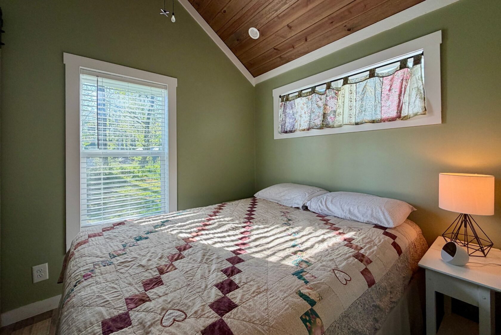 Cozy bedroom at 106 Meandering Lane with green walls, a patterned quilt, pillows, lamp, and decorative curtain.