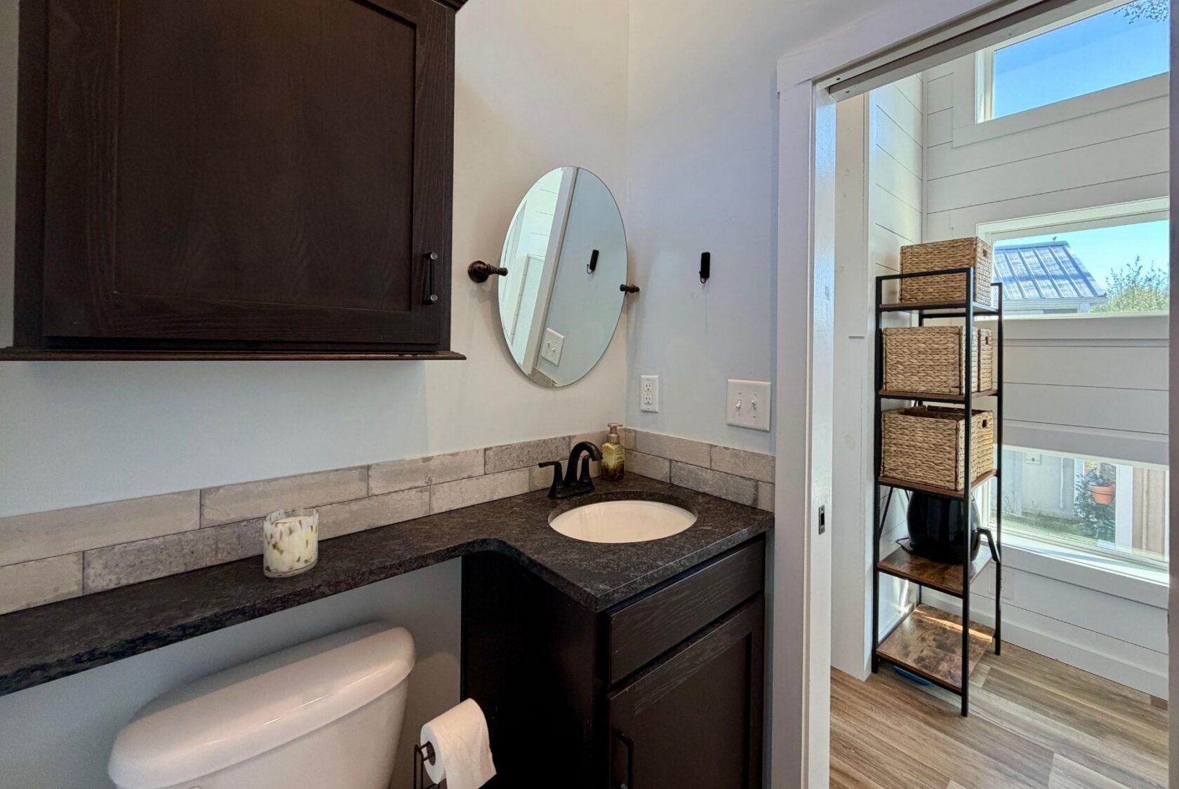 Modern bathroom at 106 Meandering Lane with dark cabinets, round mirror, and open shelving in a sunlit adjacent room.