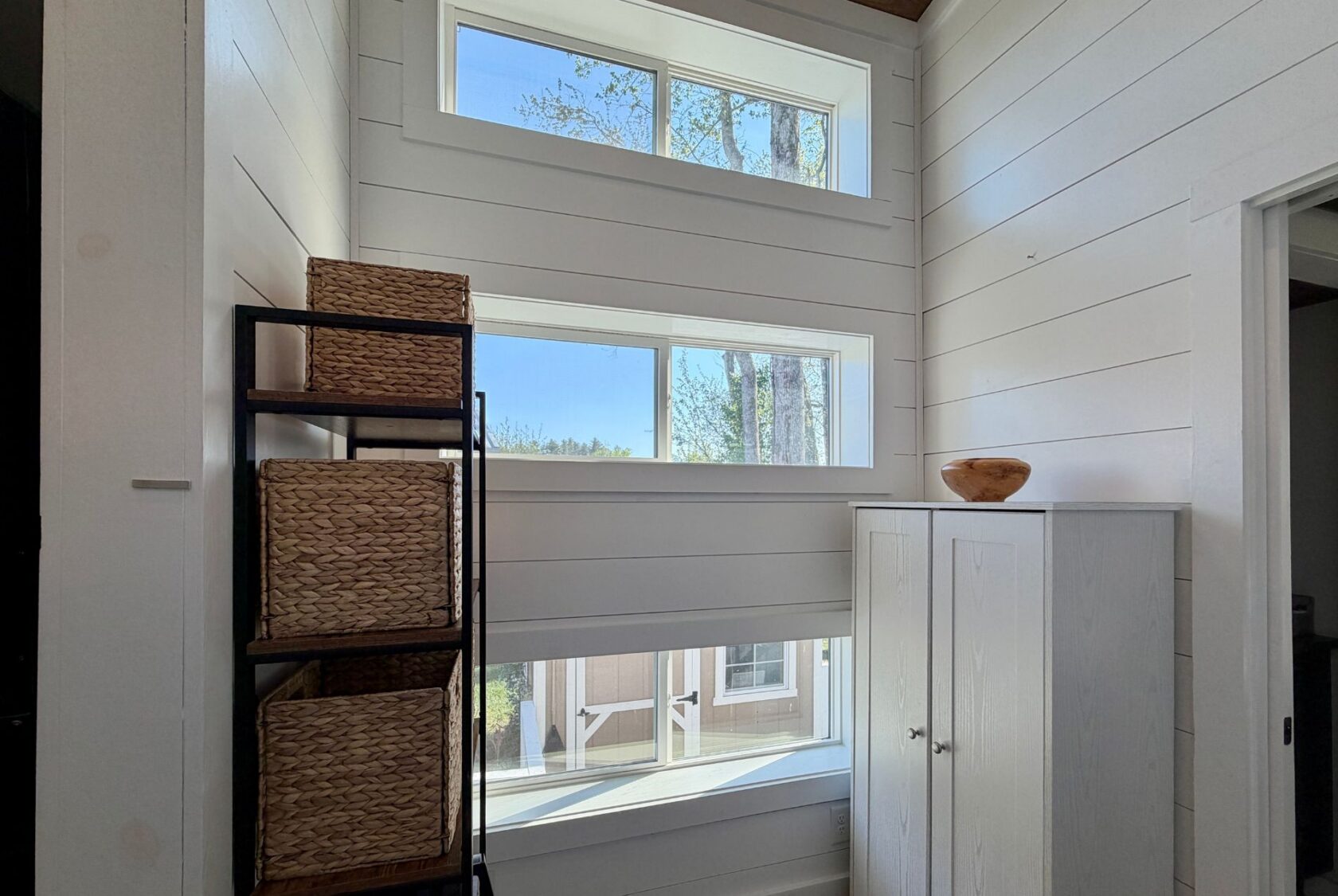 Sunny corner at 106 Meandering Lane with white shiplap walls, three windows, wicker baskets, and a small white cabinet.