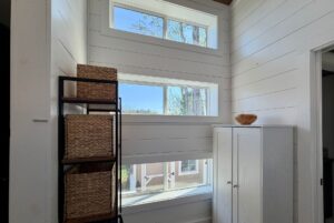 Sunny corner at 106 Meandering Lane with white shiplap walls, three windows, wicker baskets, and a small white cabinet.