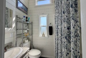 Small white bathroom on Drifting Brook Lane with patterned shower curtain, shelves above the toilet, and natural light.