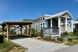 Gray tiny house with a porch and blue curtains on Strolling Easy Lane, beside a wooden carport and gravel driveway.