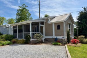 A small beige house at 59 Drifting Brook with a screened porch and gravel driveway, surrounded by greenery and blooming bushes.