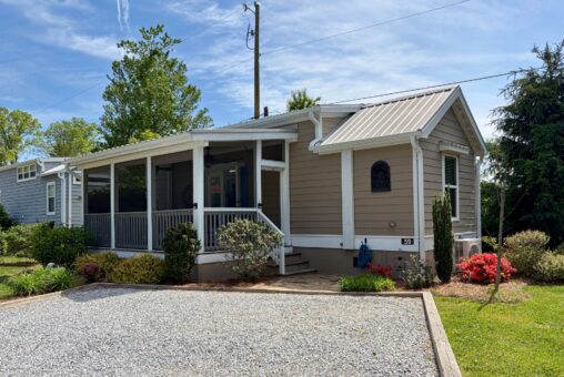 A small beige house at 59 Drifting Brook with a screened porch and gravel driveway, surrounded by greenery and blooming bushes.