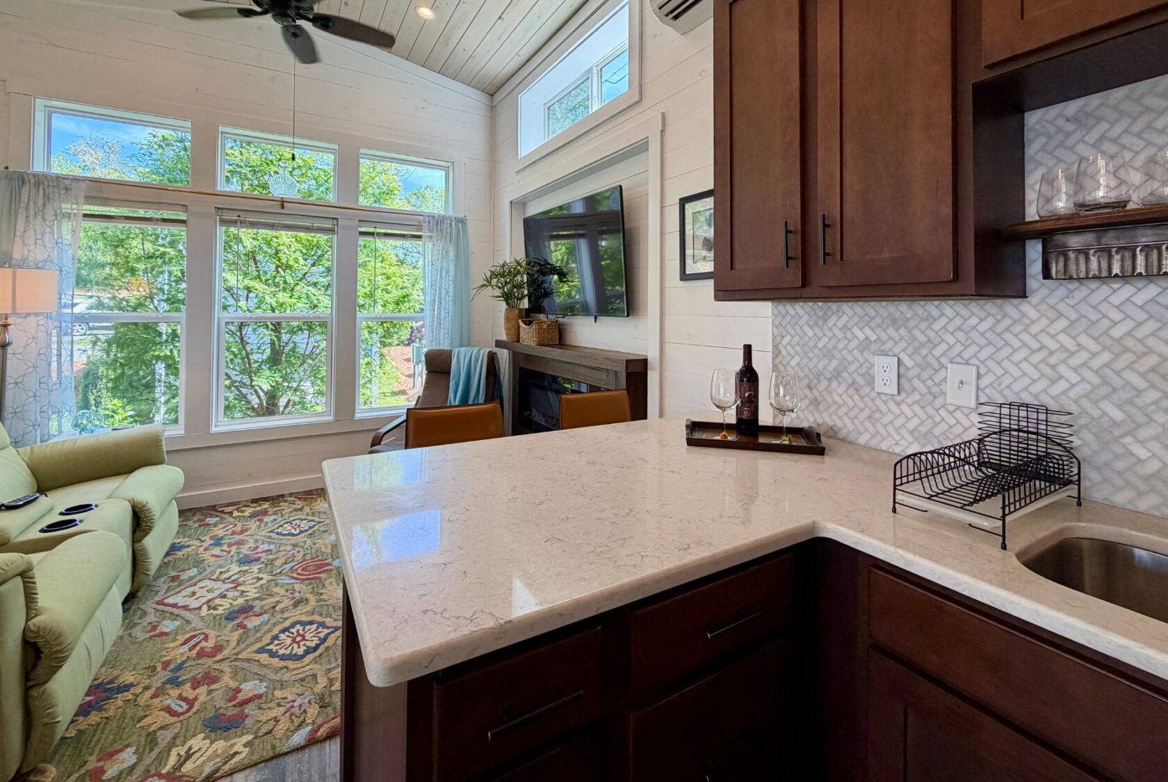 A cozy living room and kitchen at 59 Drifting Brook Lane features large windows, a green sofa, TV, and wine bottle on the counter.