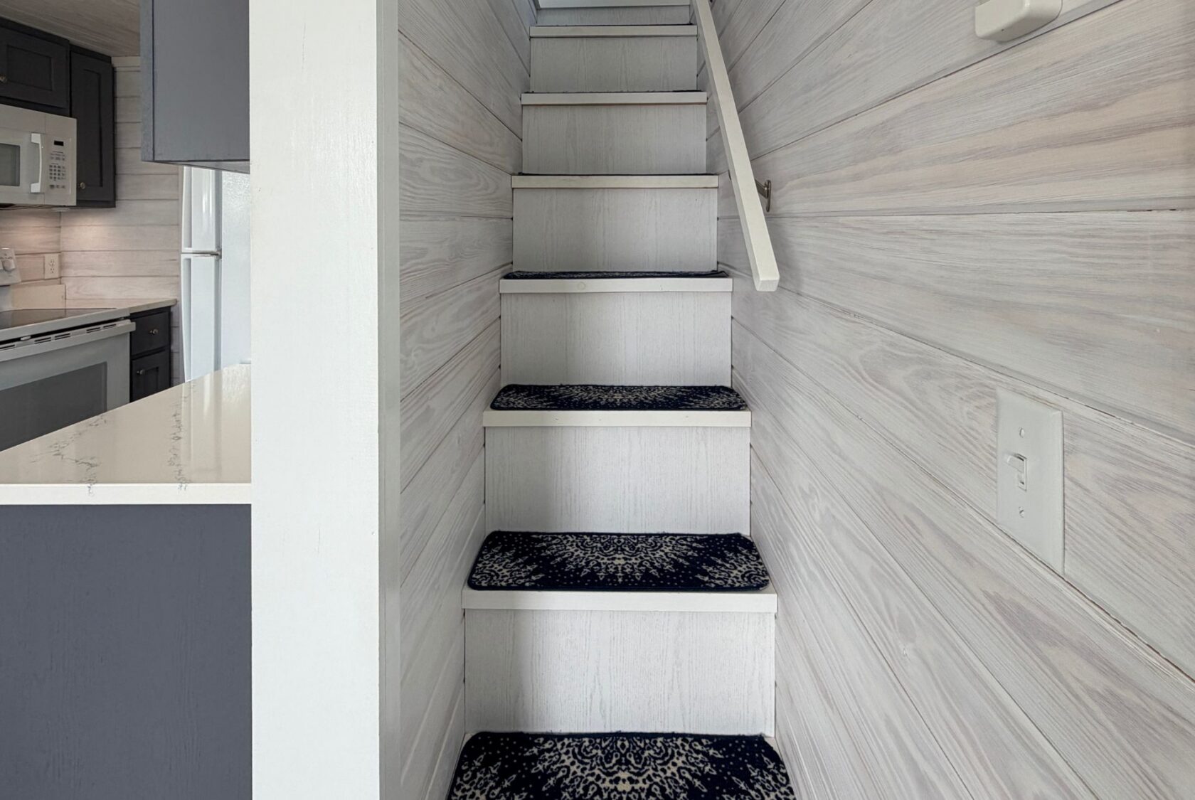 Narrow staircase with carpet runners and white wood walls on 15 Easy Lane, strolling next to a kitchen area.