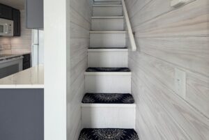 Narrow staircase with carpet runners and white wood walls on 15 Easy Lane, strolling next to a kitchen area.