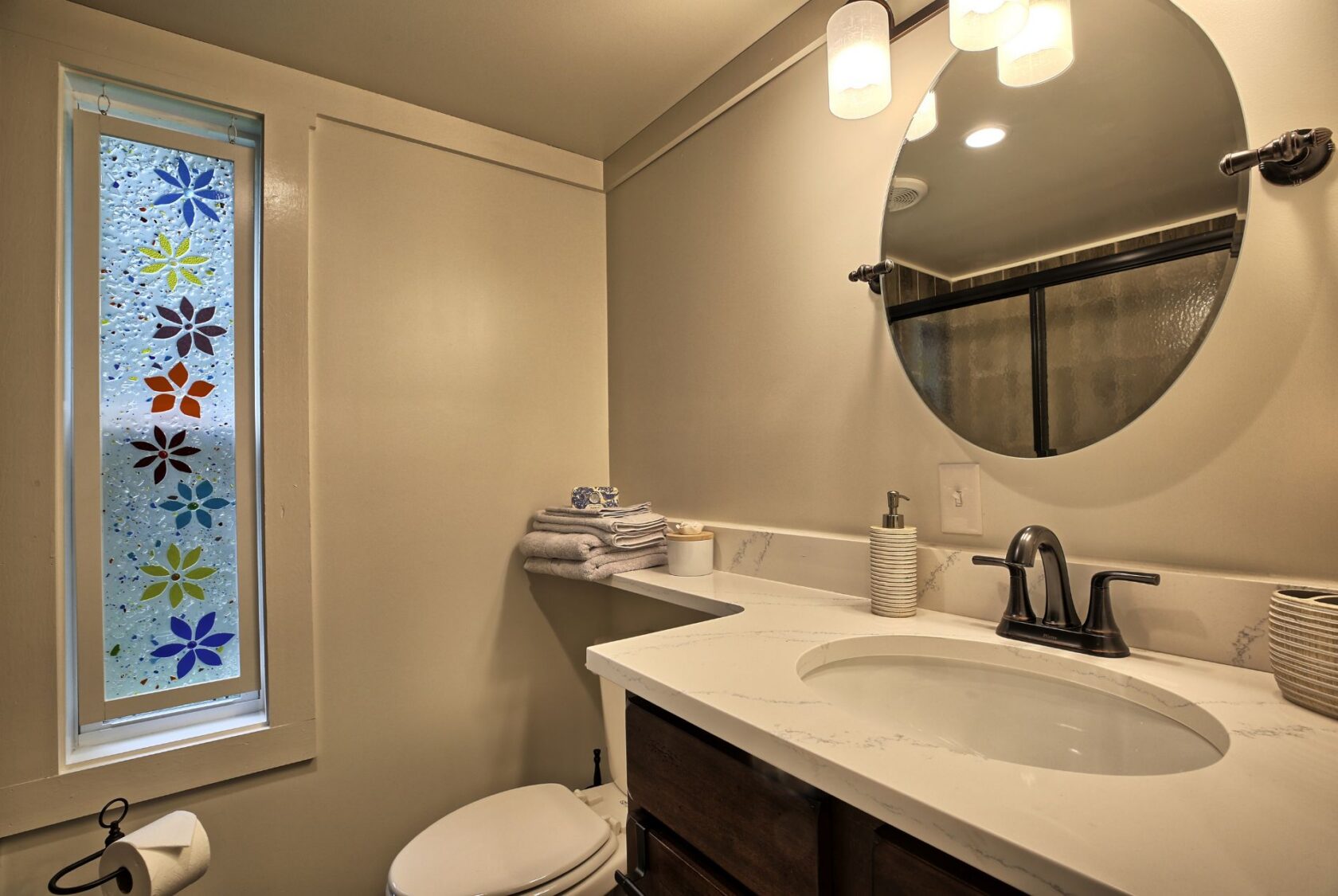 Modern bathroom at 132 Drifting Brook with round mirror, marble sink, stacked towels, and floral window—ideal real estate.