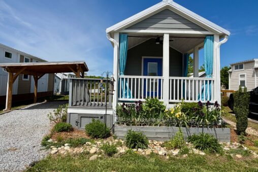 Small gray tiny house on Strolling Easy Lane with a front porch, blue curtains, and a flower-filled garden.
