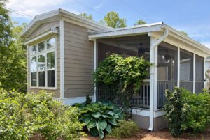 A screened porch and large windows grace this tan home at 59 Drifting Brook, surrounded by green plants and shrubs.