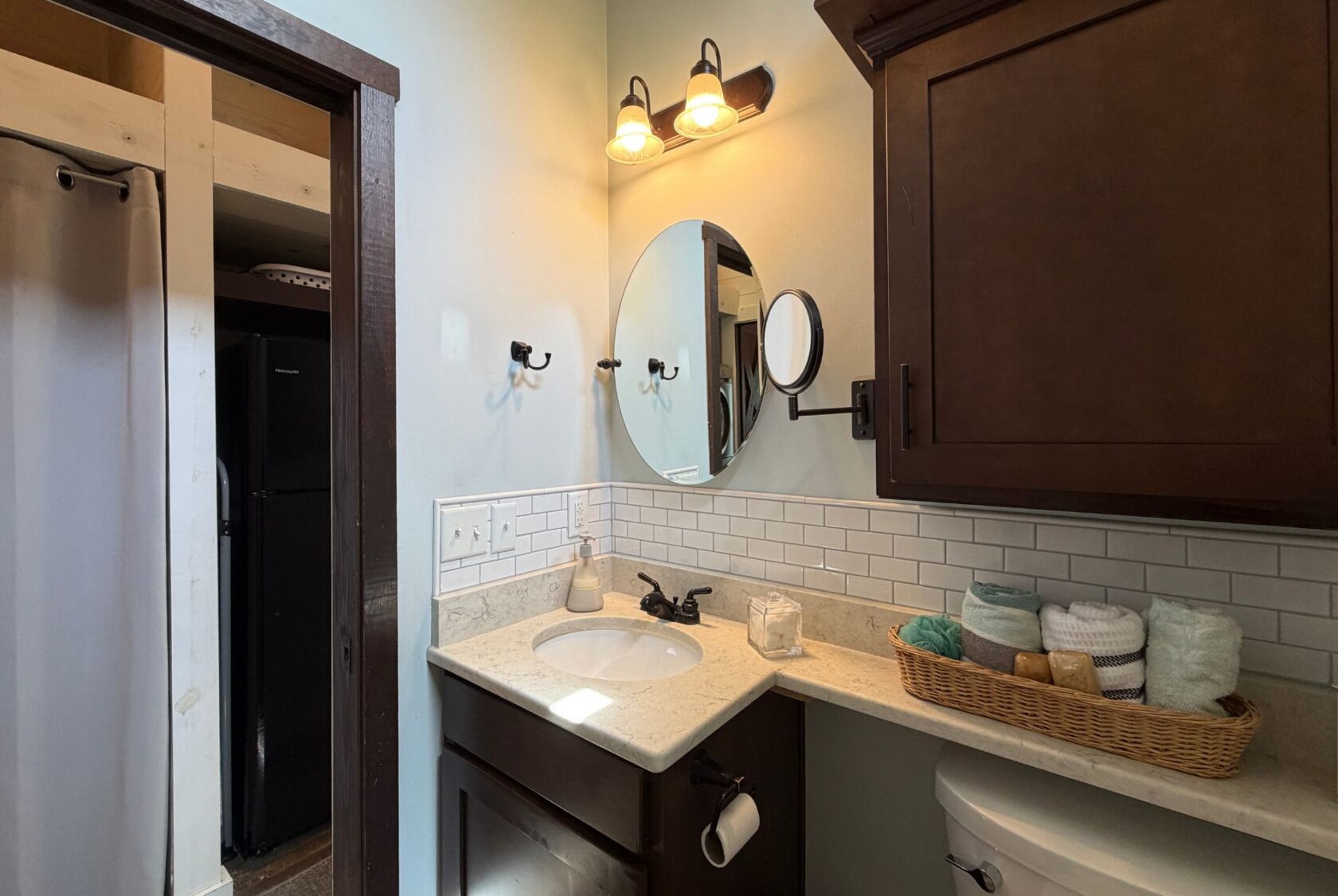 Small bathroom at 59 Drifting Brook Lane features dark wood cabinets, round mirror, and white subway tile backsplash.