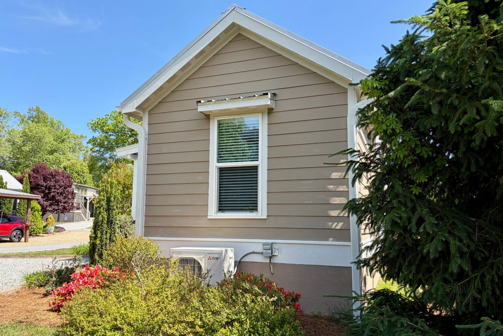 Side of a beige house at 59 Drifting Brook, with a window, flowers, bushes, and trees on a sunny day.