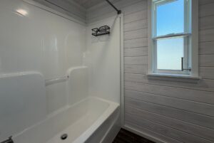 White bathtub and shower with wall shelf next to a window in this Strolling Easy Lane wood-paneled bathroom.