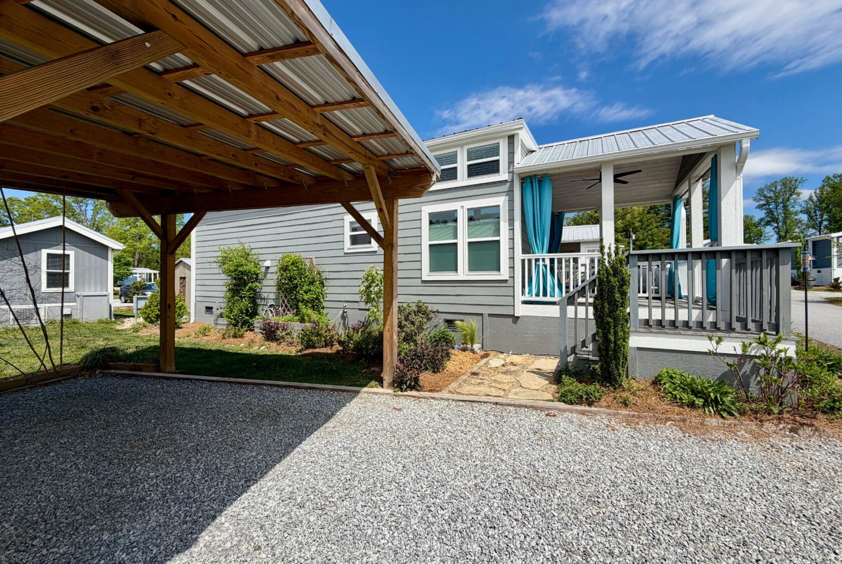 A small gray house with a porch sits on Strolling Easy Lane, next to a wooden carport on a gravel driveway.