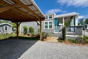 A small gray house with a porch sits on Strolling Easy Lane, next to a wooden carport on a gravel driveway.