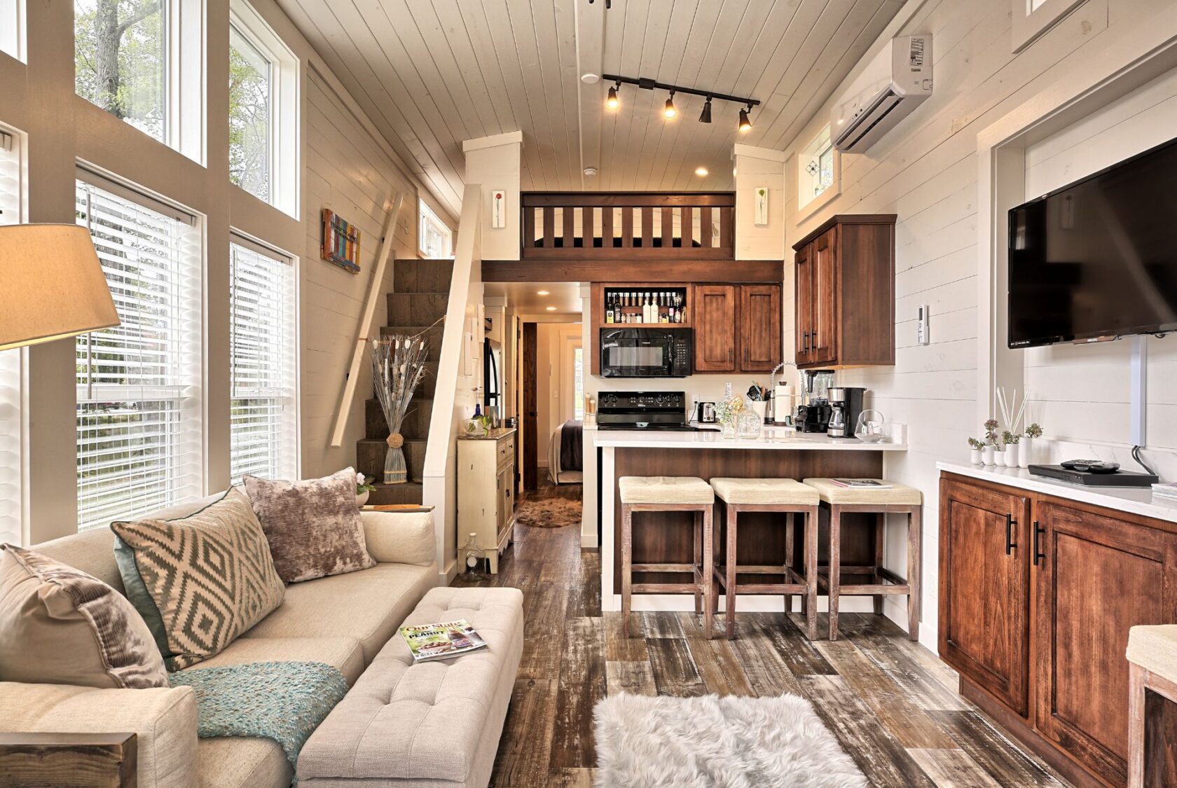 Modern tiny house interior at 132 Drifting Brook Lane with loft, cozy couch, kitchen, bar stools, and bright windows.