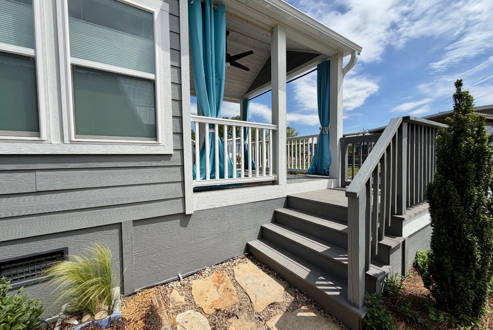Gray house on Strolling Easy Lane with a porch, blue curtains, stairs, plants, and stones under a sunny sky.