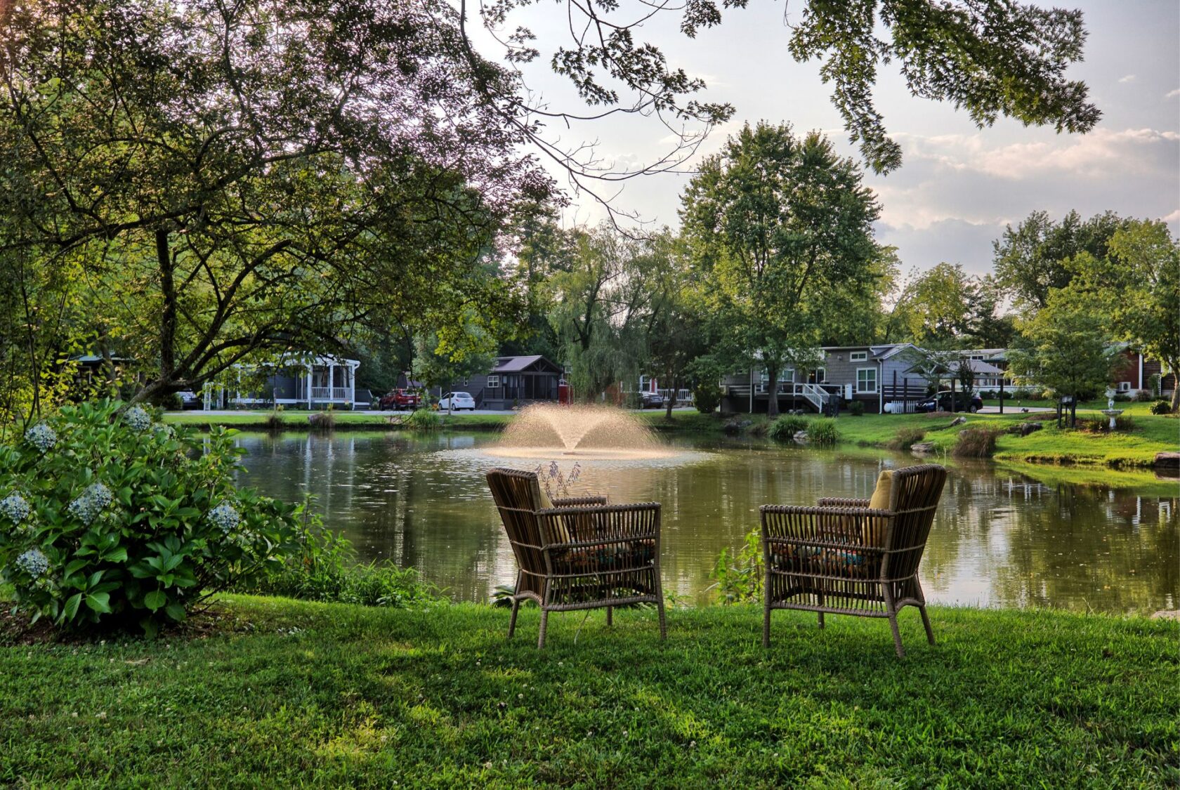 Two wicker chairs face a pond with fountain at 132 Drifting Brook, surrounded by trees, grass, and homes on a sunny day.