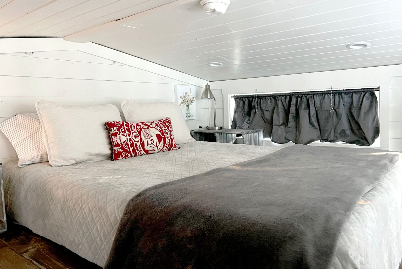 Cozy bedroom at 132 Drifting Brook, featuring a large bed, gray blanket, red pillow, lamp, flowers, and curtained window.