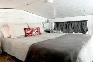 Cozy bedroom at 132 Drifting Brook, featuring a large bed, gray blanket, red pillow, lamp, flowers, and curtained window.
