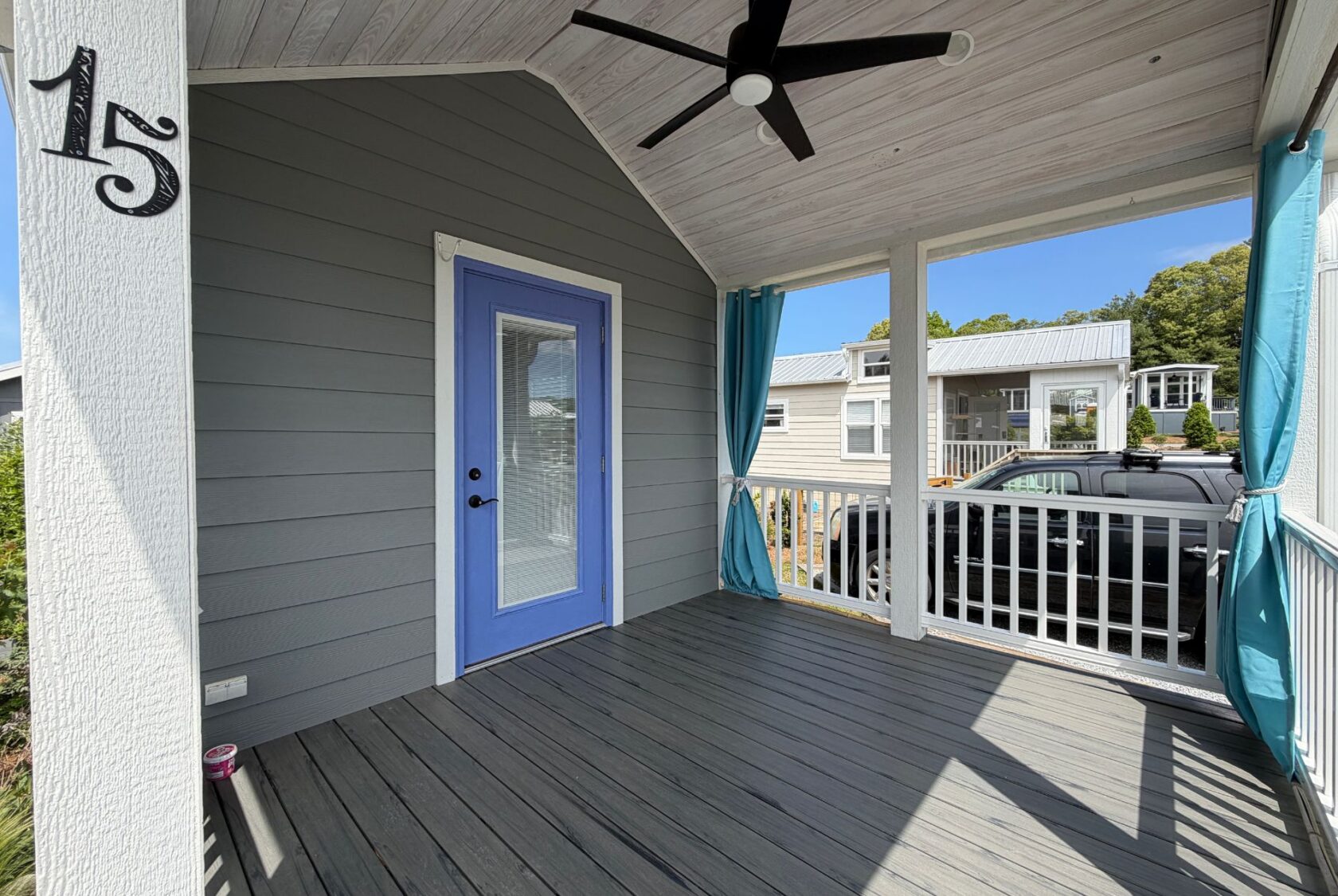 Covered porch on Strolling Easy Lane with gray siding, blue door, number 15, ceiling fan, and blue-curtained white railing.