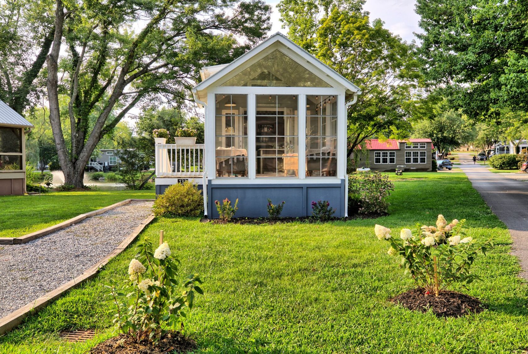A tiny house at 132 Drifting Brook Lane with a screened porch and garden, surrounded by green grass and trees.