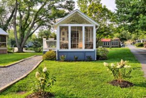 A tiny house at 132 Drifting Brook Lane with a screened porch and garden, surrounded by green grass and trees.