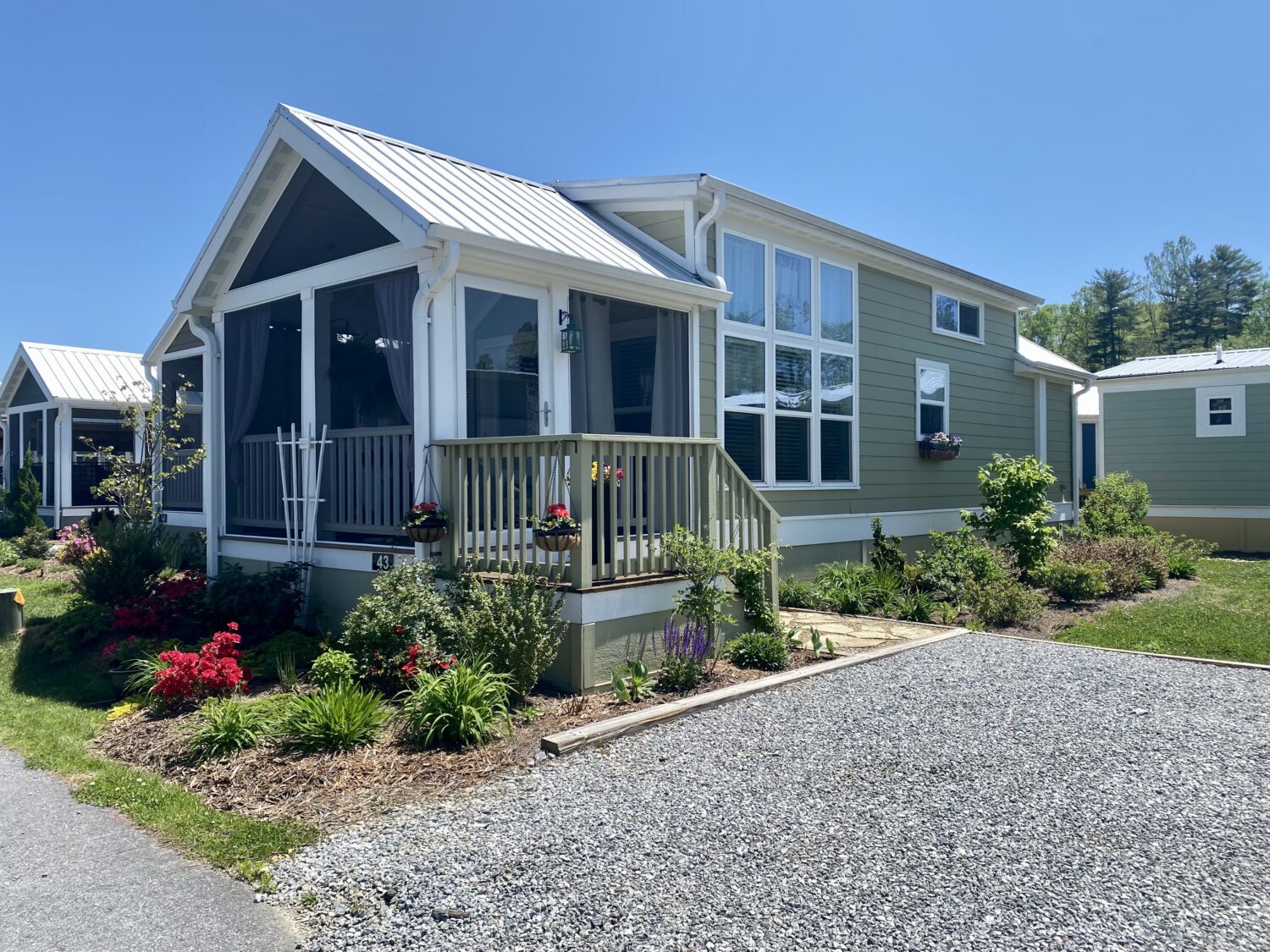 A small, modern cottage with a porch on Skipping Stone Lane, surrounded by flowers and greenery under a clear blue sky.