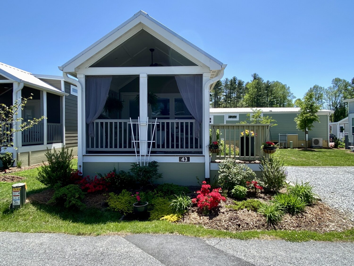 Charming small white cottage on Skipping Stone Lane, with a front porch amid colorful flowers and sunny greenery.