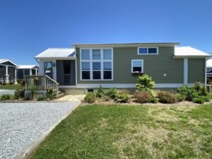 A small, modern green house on 43 Skipping Stone Lane with a porch and manicured lawn under a clear blue sky.