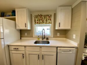 White kitchen cabinets, a sink under a window with floral curtains, and a dishwasher on Skipping Stone Lane.