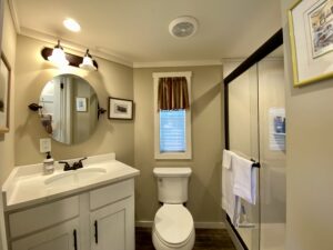 Charming small bathroom on Skipping Stone Lane with round mirror, sink, toilet, window, and glass shower with towel.